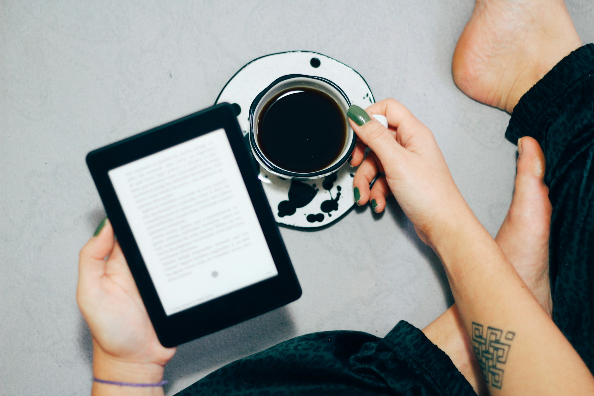 Overhead view of a person with green fingernails sitting on the floor, holding a black and white cup of coffee in one hand and an e-reader in another.