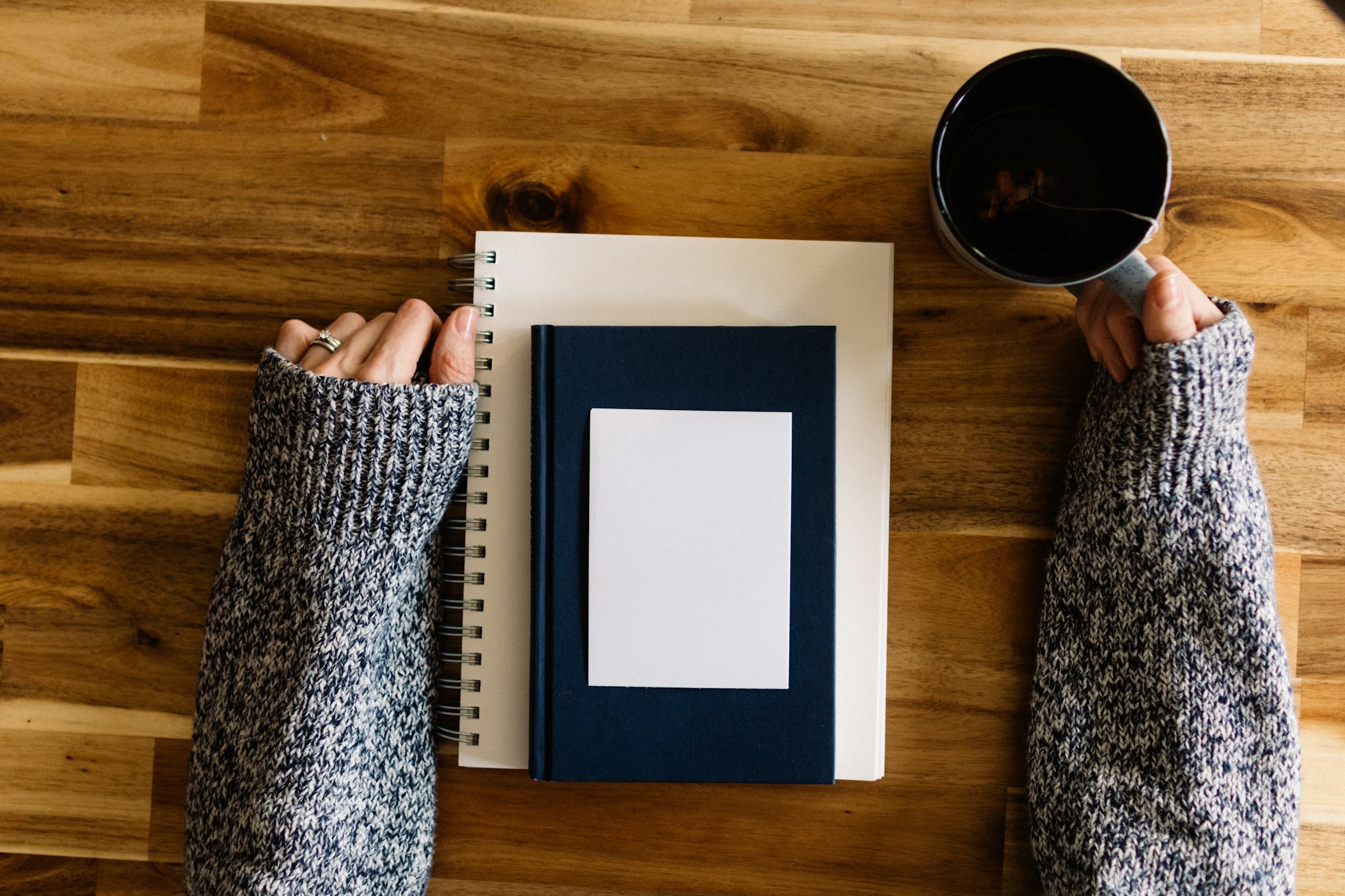 A person's arms rest on a table top, a cup of coffee in one hand. Between their arms is a stack of notebooks, each smaller than the one beneath it.