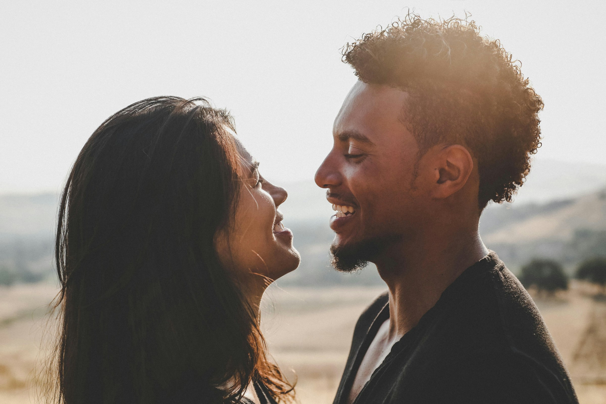 A couple stands face to face on a beach, smiling at one another.