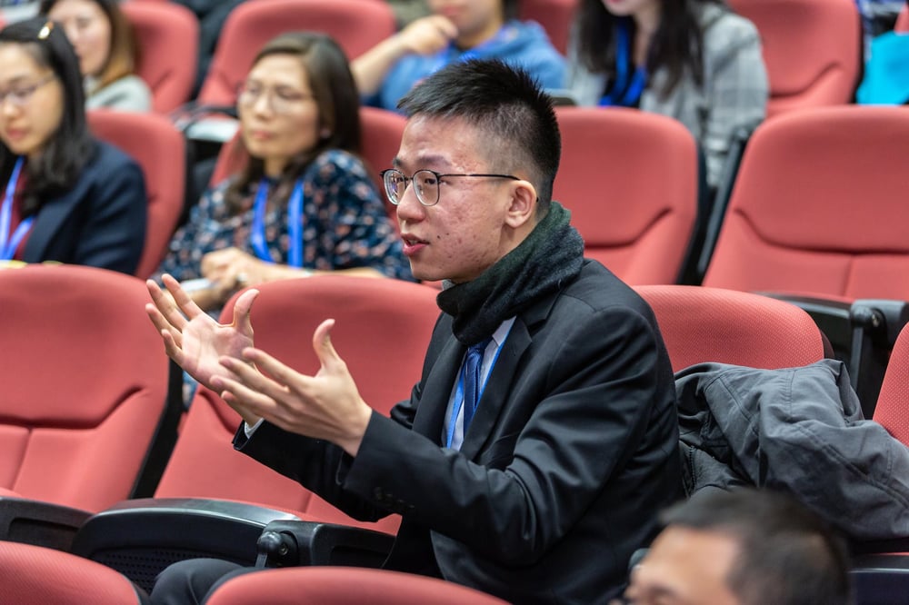A person gestures while speaking from their seat in an auditorium.
