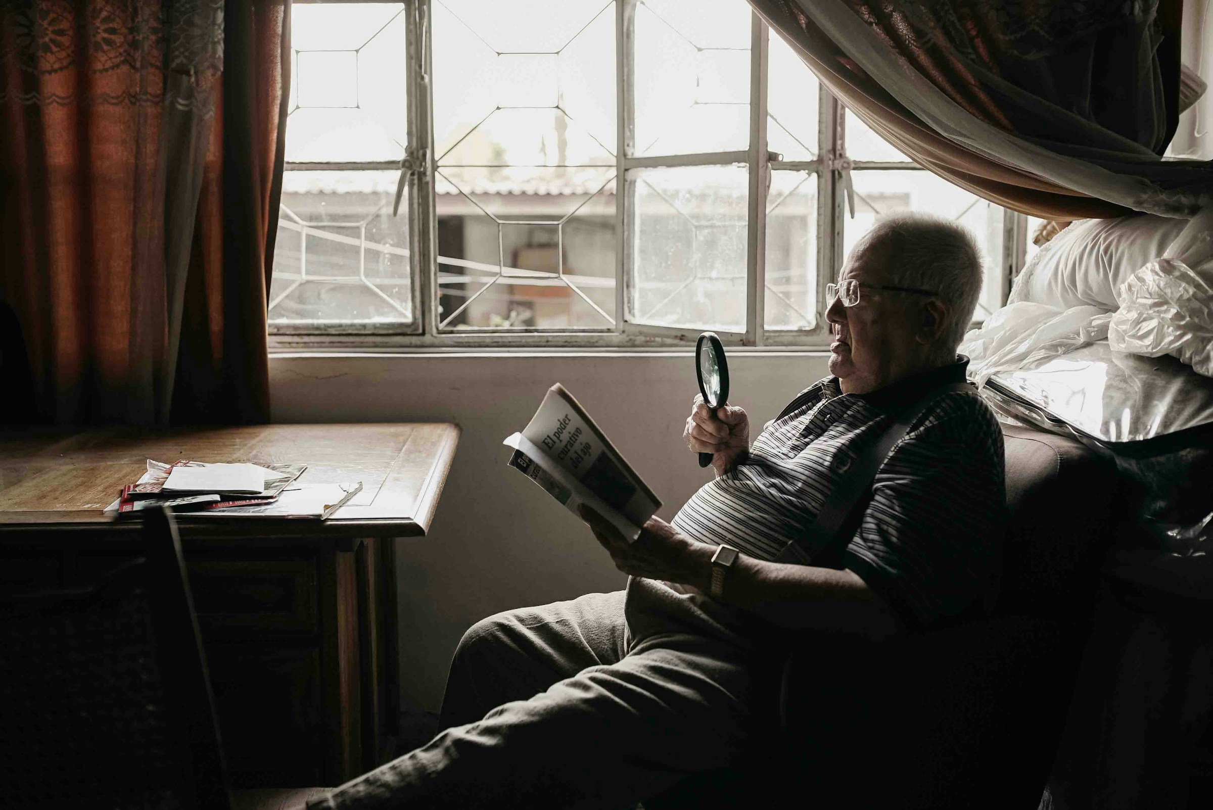 A smiling person sits by a window, reading a book with a magnifying glass.