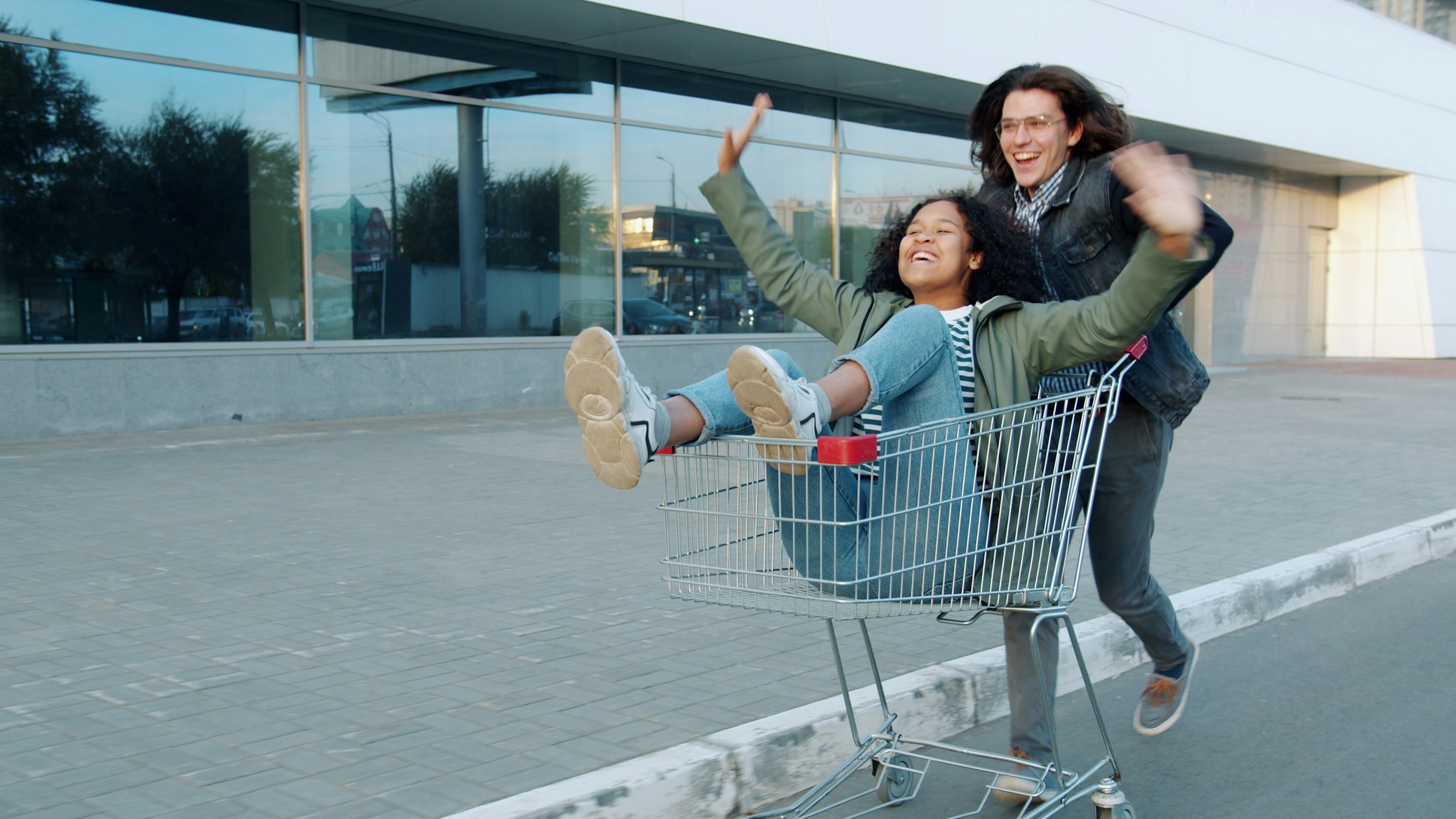 A person with shoulder-length hair and glasses pushes a laughing person in a shopping cart.