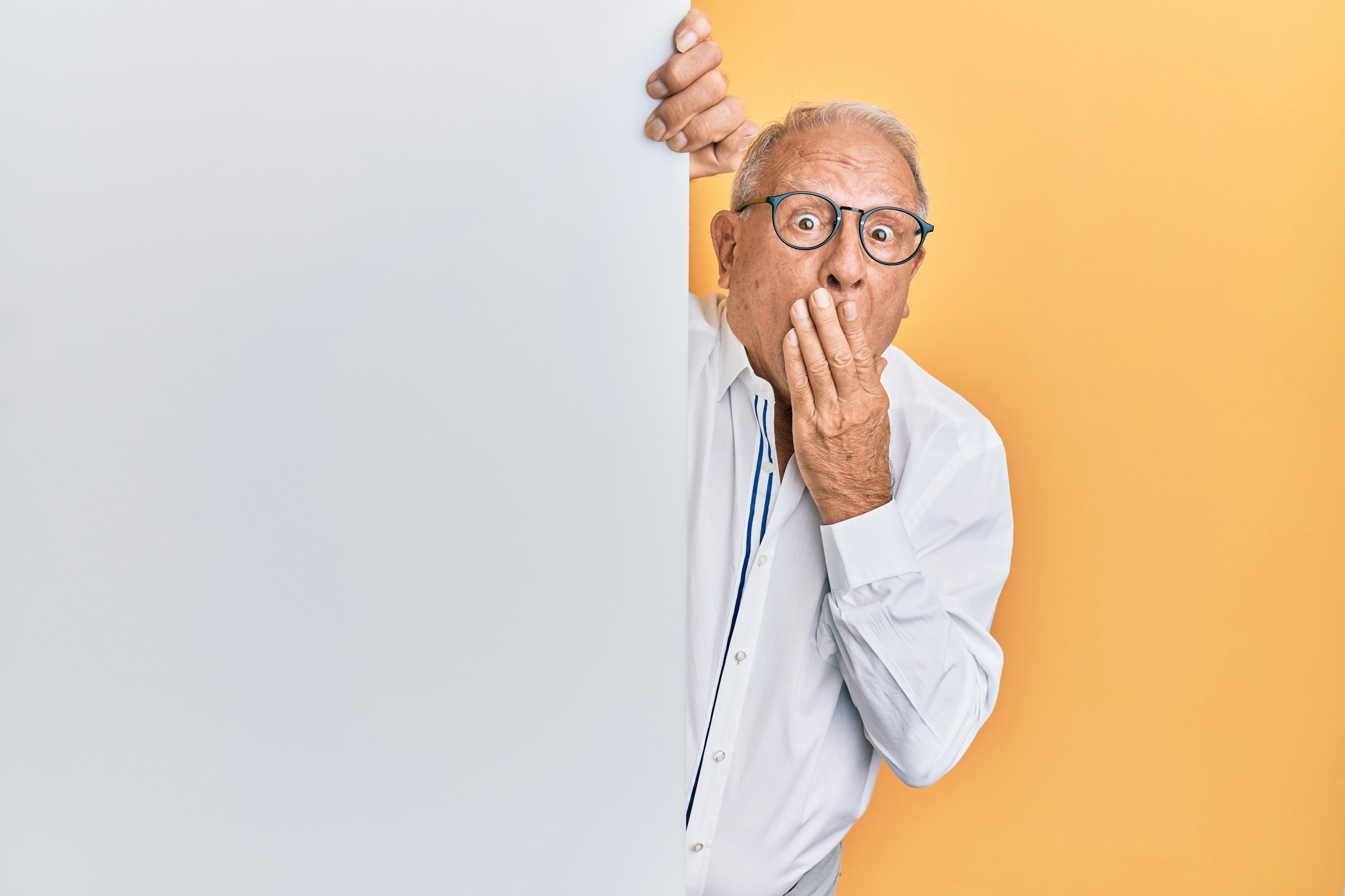 An older person with glasses peeks around a wall looking shocked with wide eyes and a hand over their mouth.
