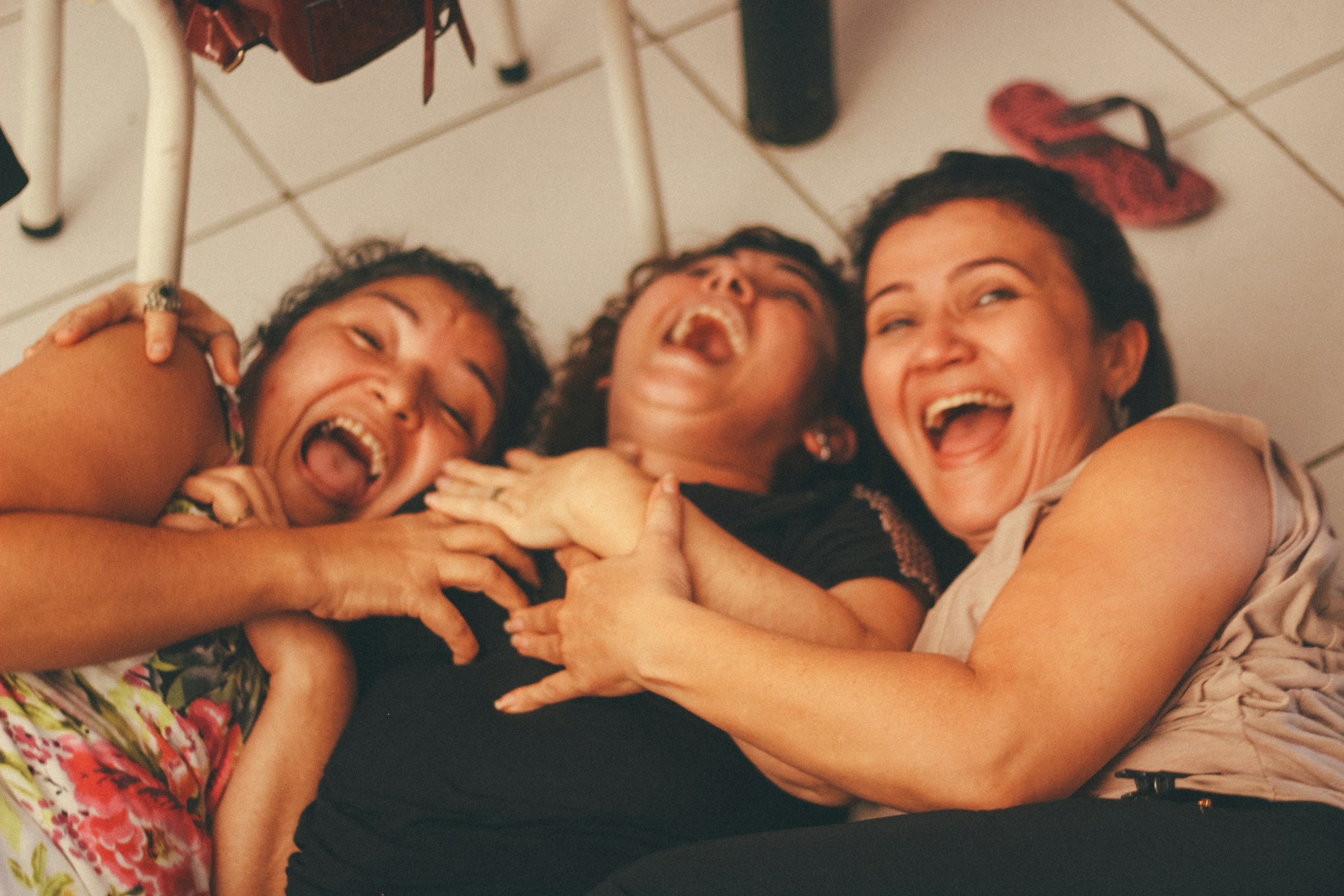 Three young adults lay on a tile floor with their arms around each other, laughing.