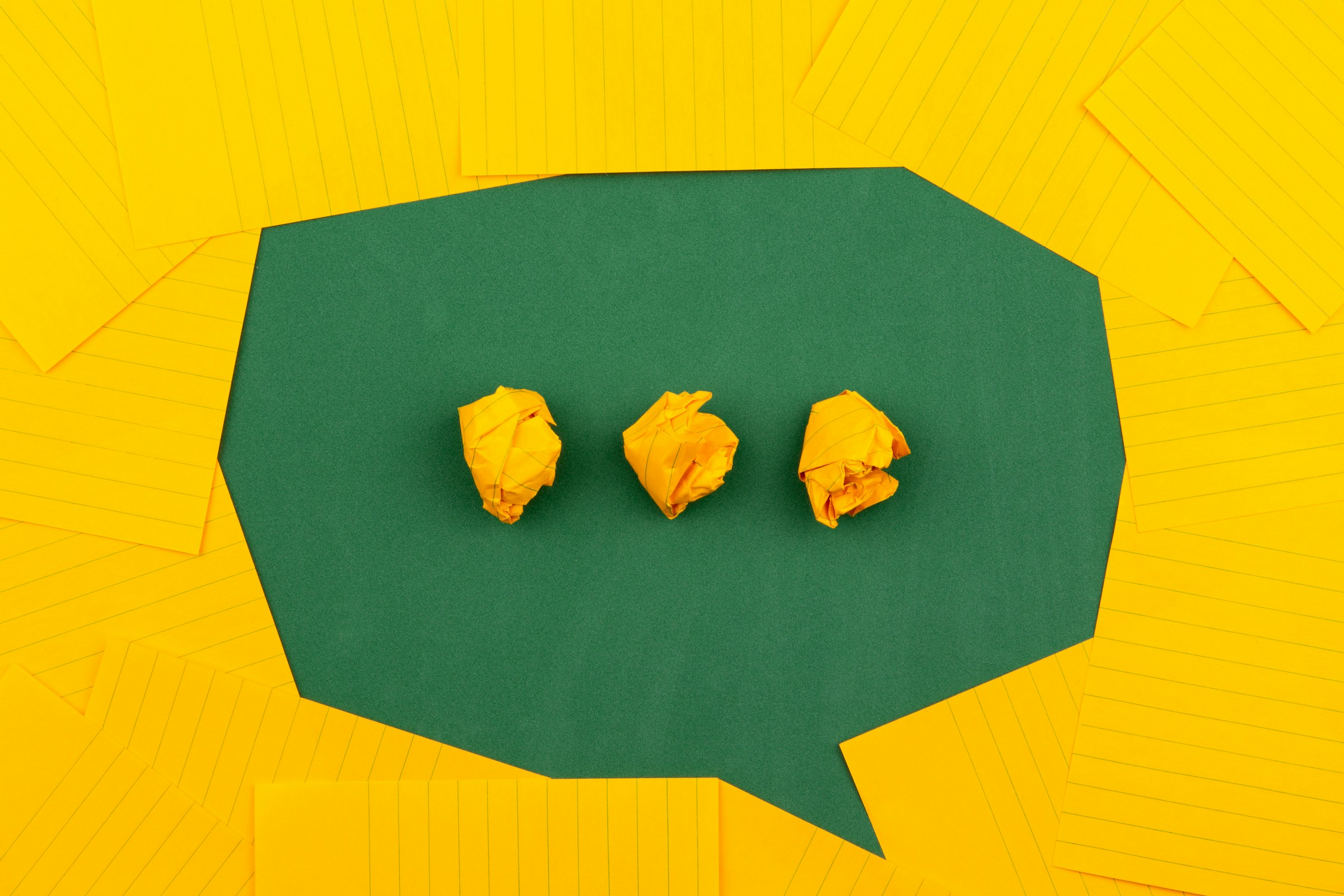 A green conversation bubble with yellow wadded up papers serving as ellipses inside of it.