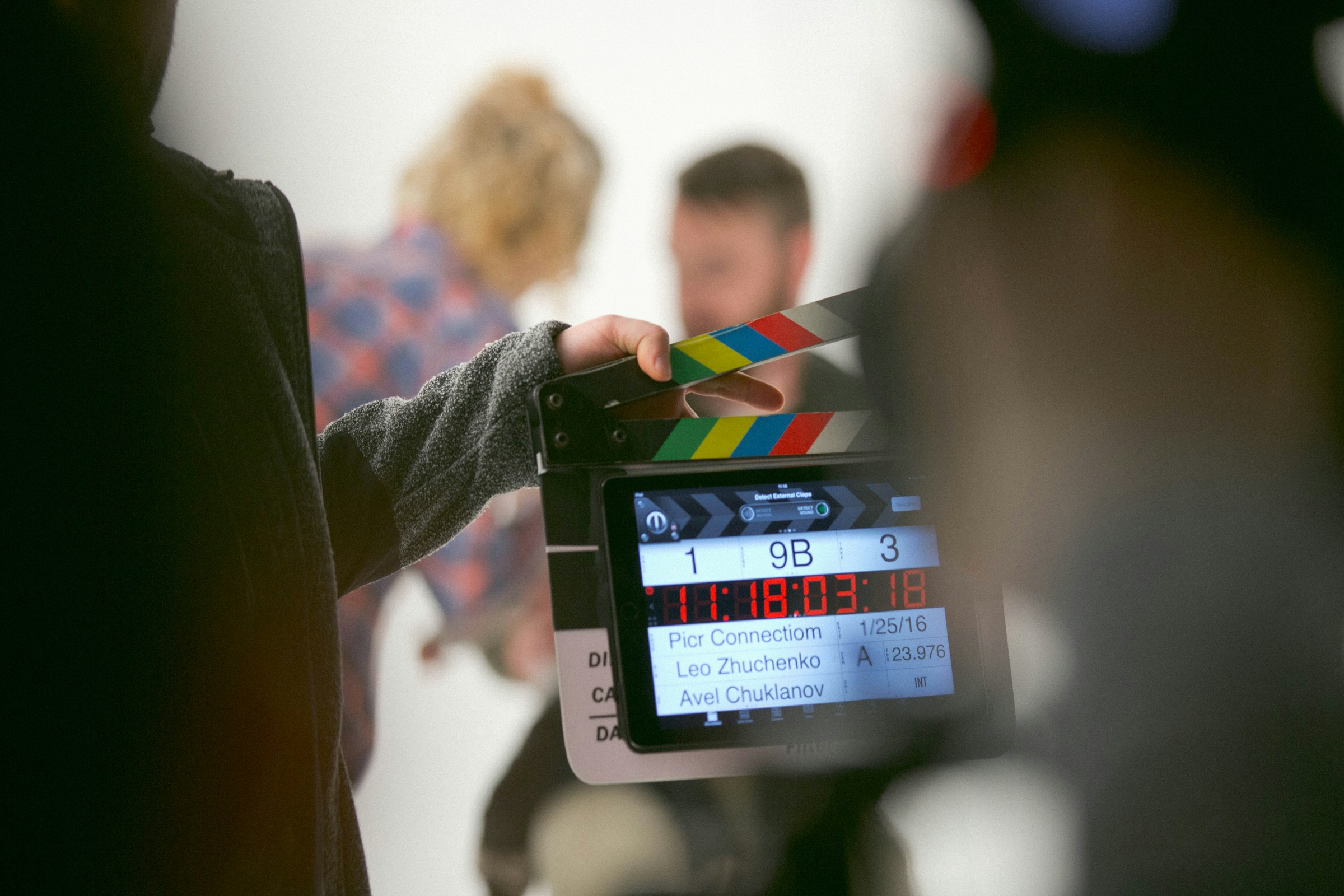 A hand holding a digital movie slate, as blurred actors talk in the background.