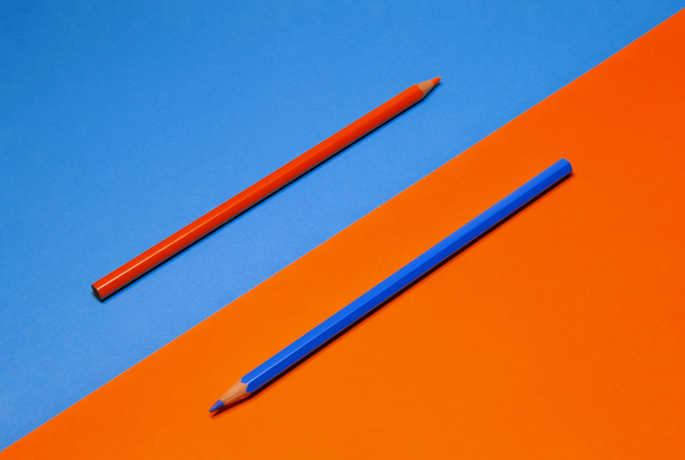 An orange pencil sits on a blue background beside a blue pencil on an orange background.